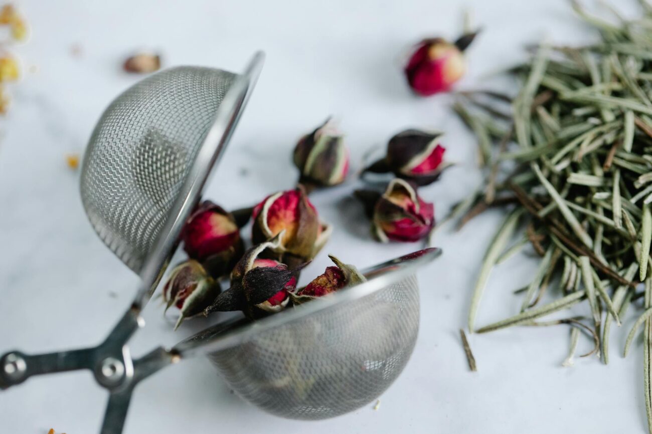 Close-up of rosebuds and herbs in a stainless steel tea strainer on a white surface, highlighting rosemary tea recipe homemade ingredients.